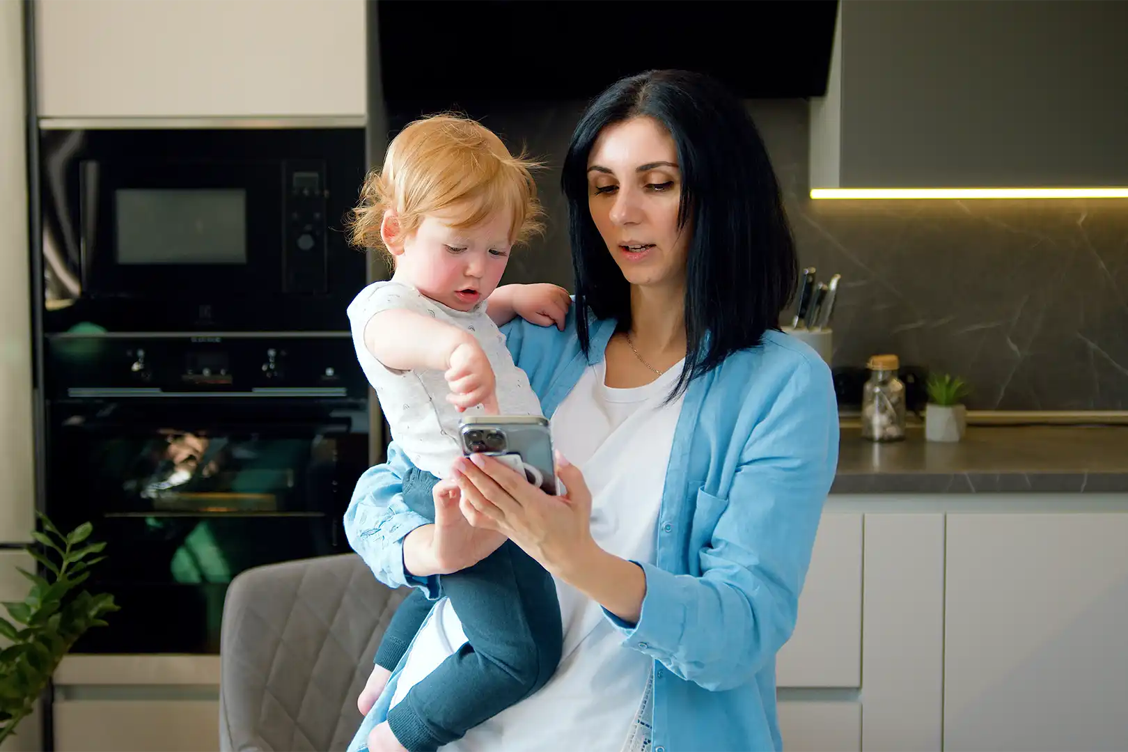 A woman stood in her kitchen cradles her toddler in one arm whilst using her phone with her other hand.